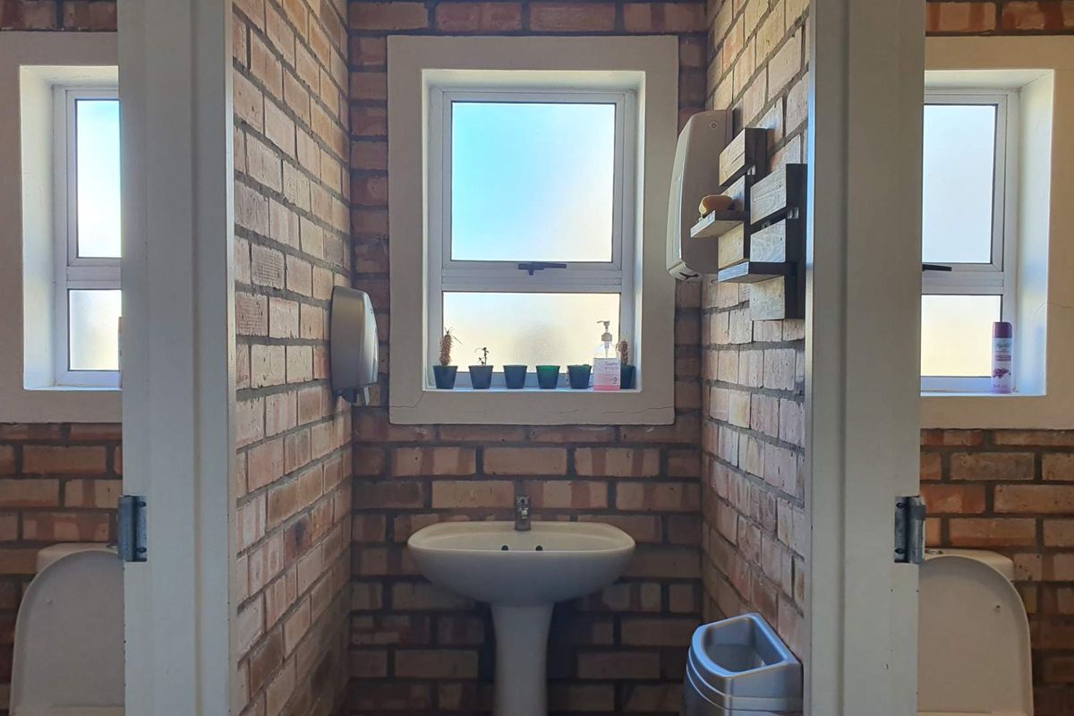 A functional bathroom featuring exposed brick walls, pedestal basin, and multiple toilet cubicles with frosted glass windows for natural light.