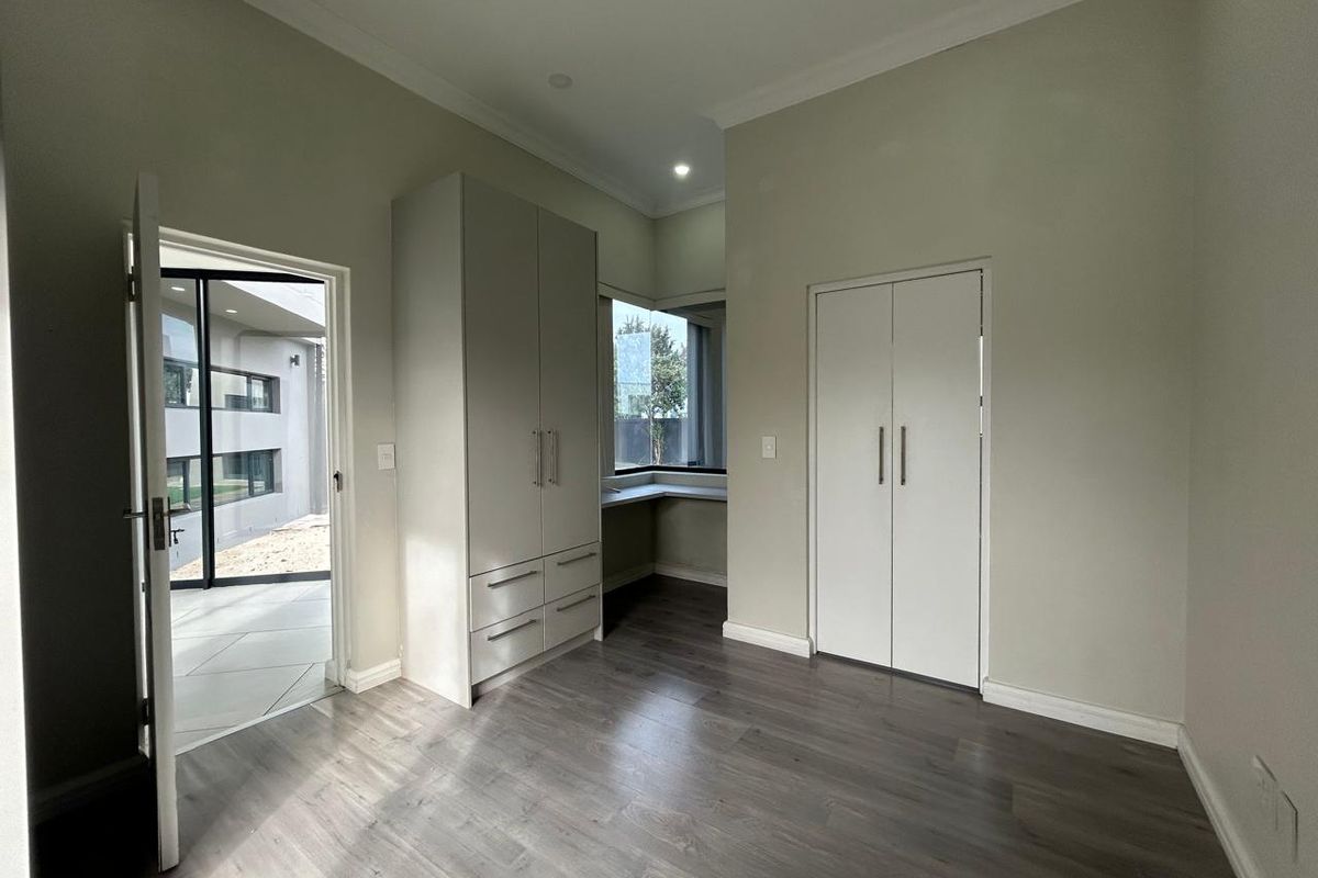 A bright, modern bedroom featuring neutral walls, laminate flooring, built-in cupboards, and a convenient corner desk area with garden access.