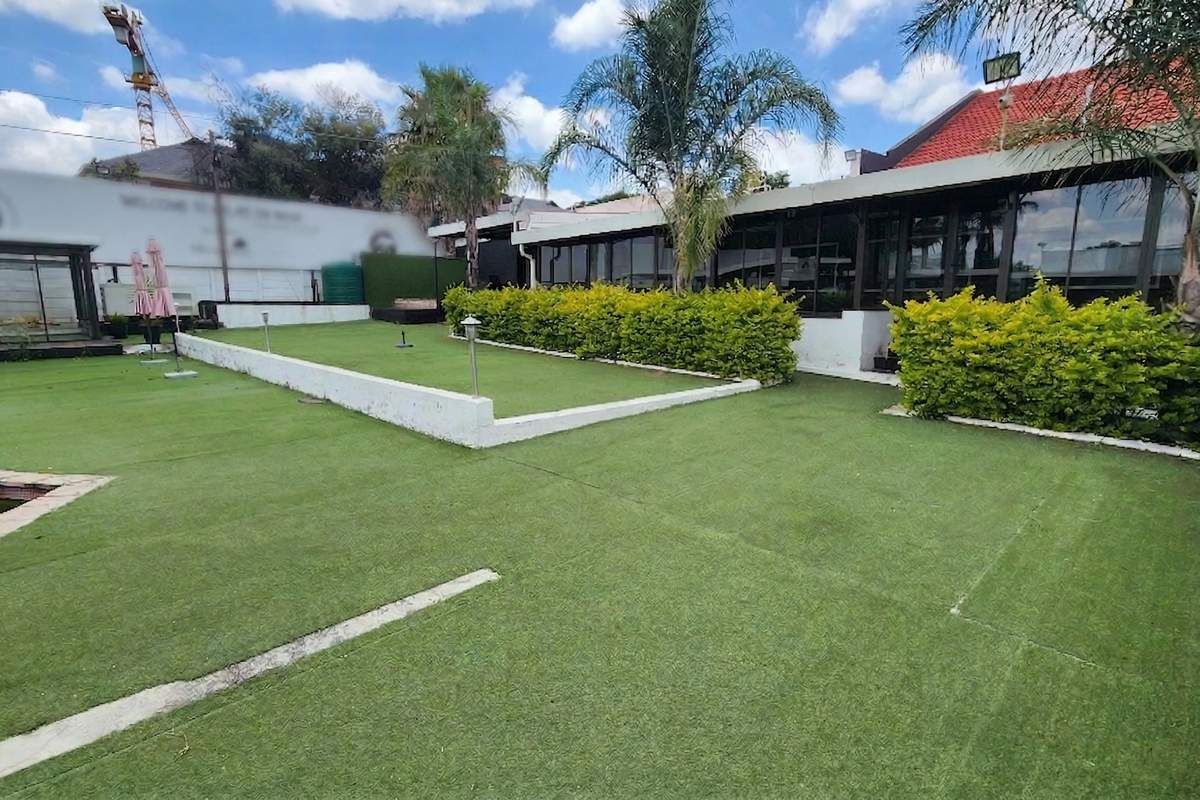 Rear exterior view featuring a large glass-fronted building, artificial turf lawn, and landscaped hedging with a retaining wall.
