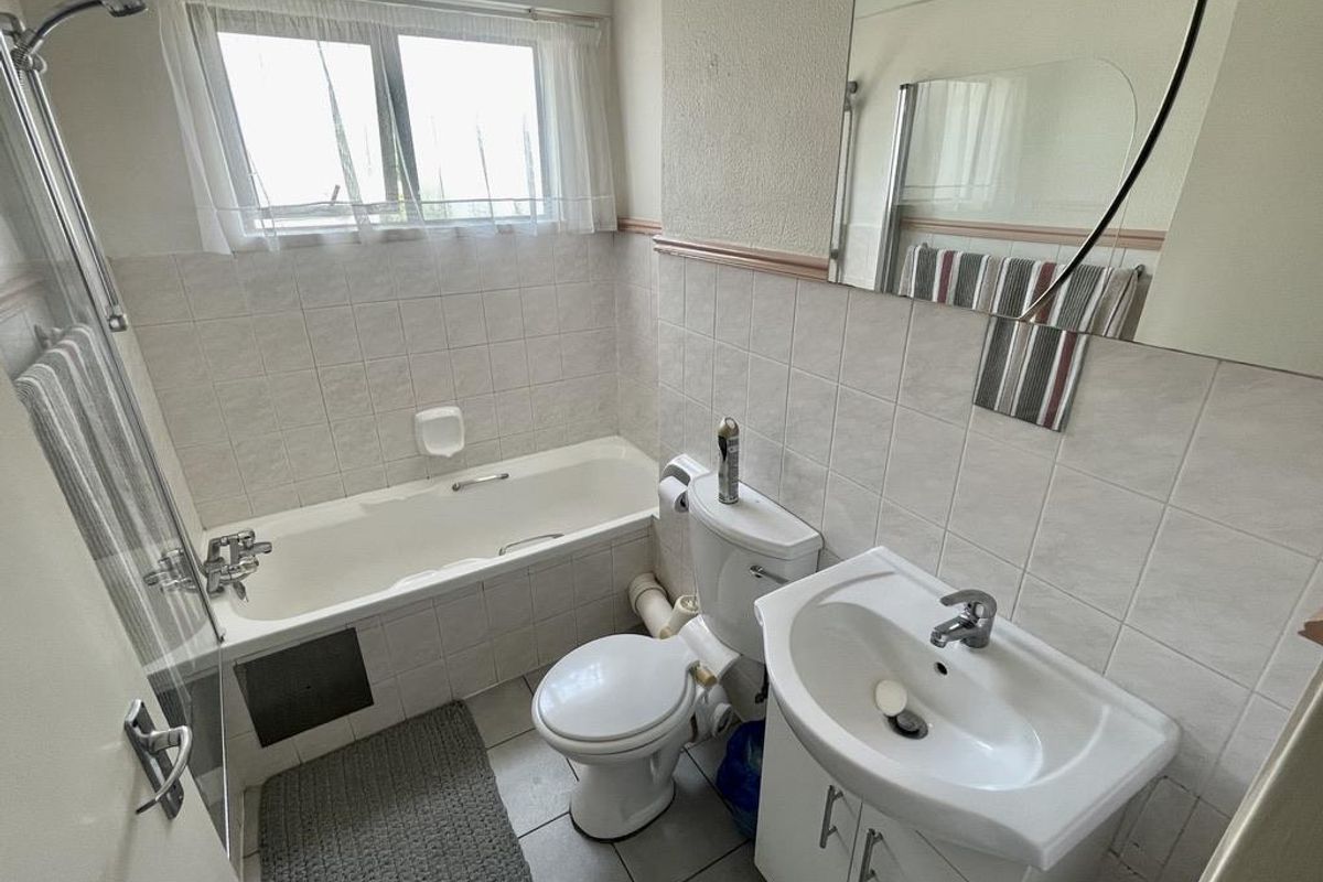 A functional bathroom featuring a bathtub with shower mixer, pedestal vanity basin, and toilet, finished with wall tiling and a window for natural light.
