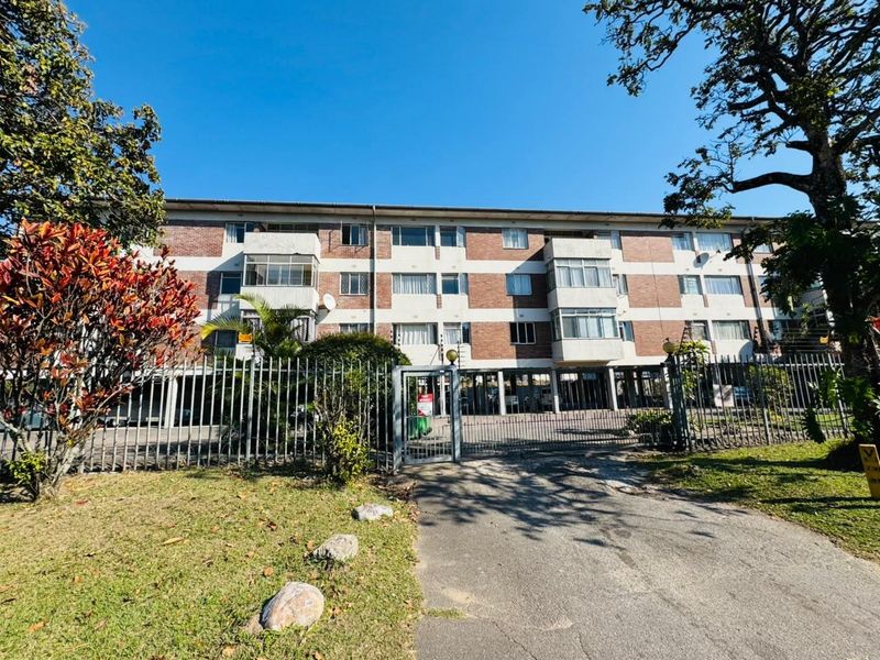 Front view of a multi-story apartment building featuring brick facade, secure gated entrance, electric fencing, and covered parking facilities.
