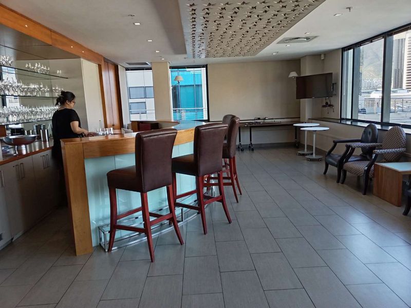 A modern office bar area featuring a built-in counter, decorative ceiling detailing, large windows with natural light, and sleek tiled flooring.