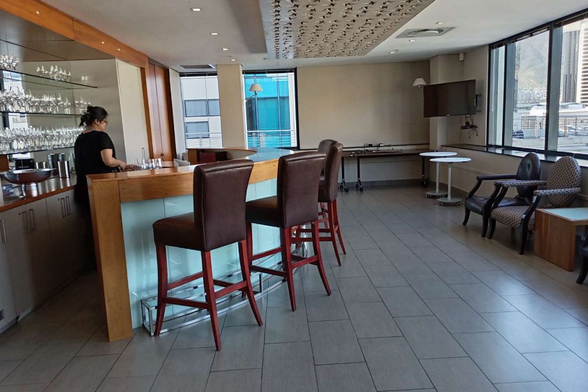 A modern office bar area featuring a built-in counter, decorative ceiling detailing, large windows with natural light, and sleek tiled flooring.