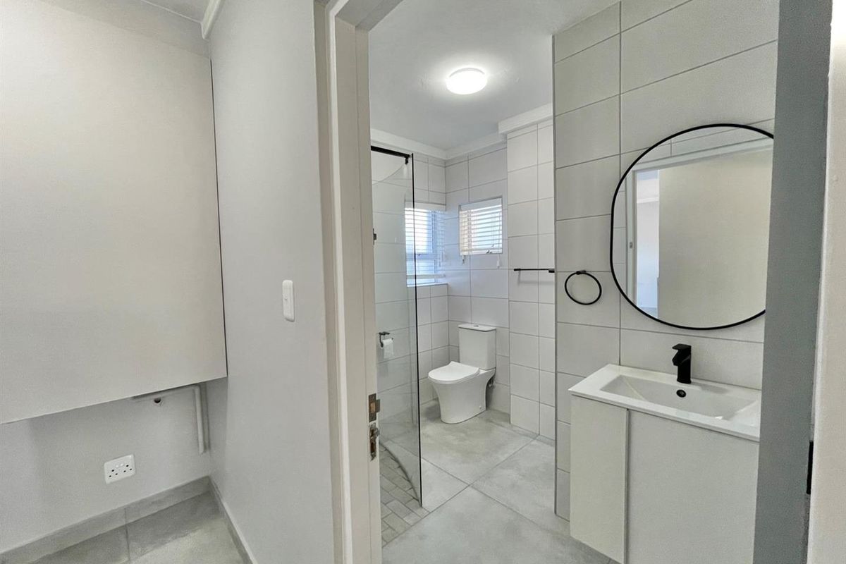 Modern bathroom featuring a glass-enclosed shower, contemporary vanity with basin, and neutral floor-to-ceiling tiling, adjacent to a utility area with plumbing connections.