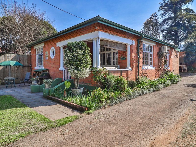 A charming single-story brick house featuring a covered front porch, paved patio area, and a long driveway set within a landscaped garden.