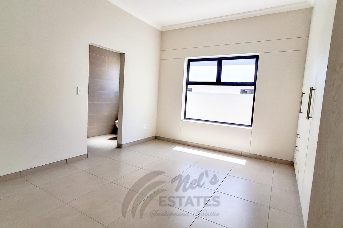 A bright, modern bedroom featuring neutral floor tiles, built-in cupboards, and an adjoining en-suite bathroom.