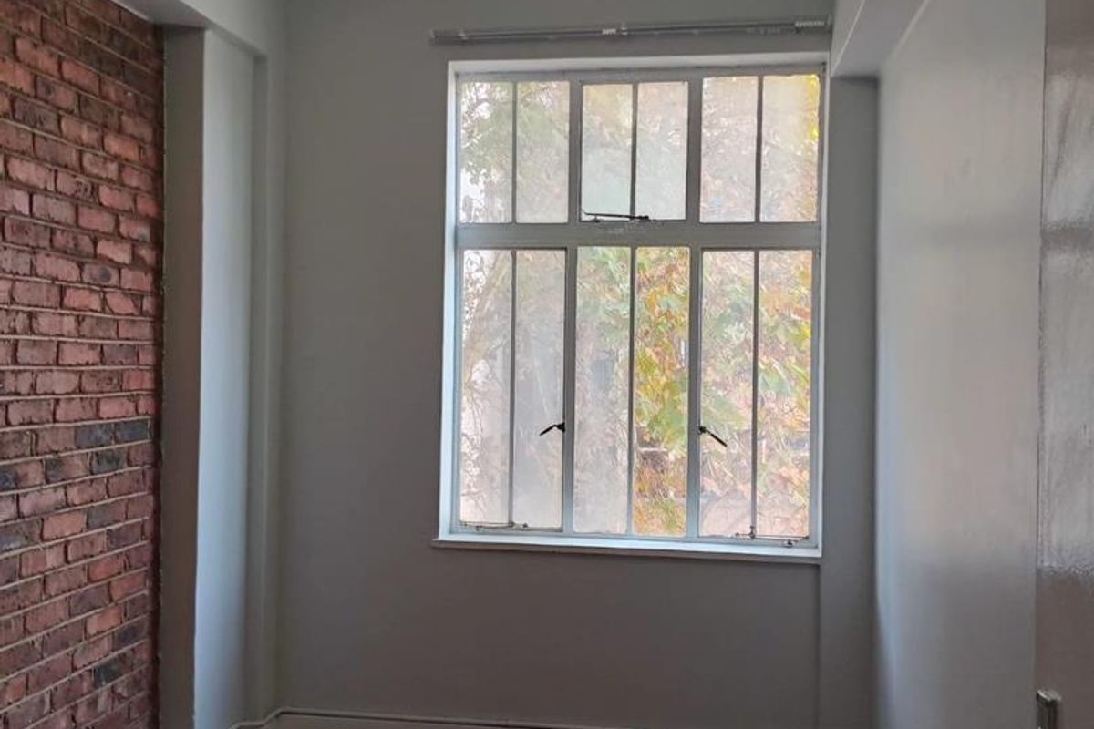 An empty bedroom featuring a characterful exposed brick feature wall, traditional parquet flooring, and a large multi-pane window allowing natural light.