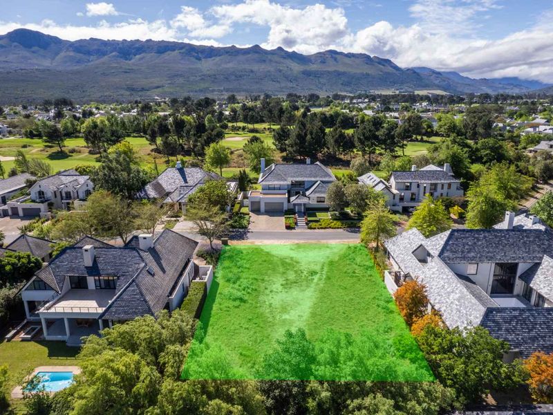 An aerial view of a vacant residential plot situated within an established estate, featuring surrounding luxury homes and scenic mountain views.
