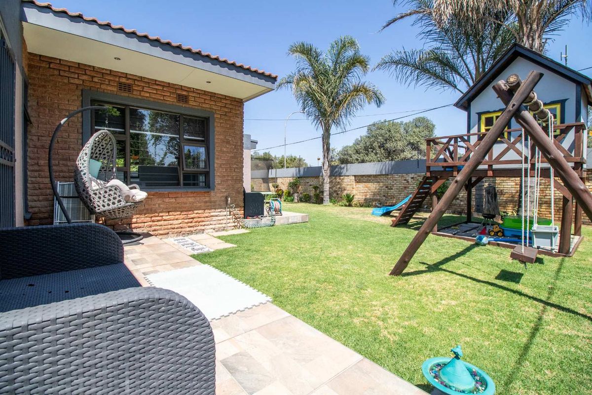 A well-maintained rear garden featuring a paved patio area, lush lawn, and a brick exterior wall with large windows.