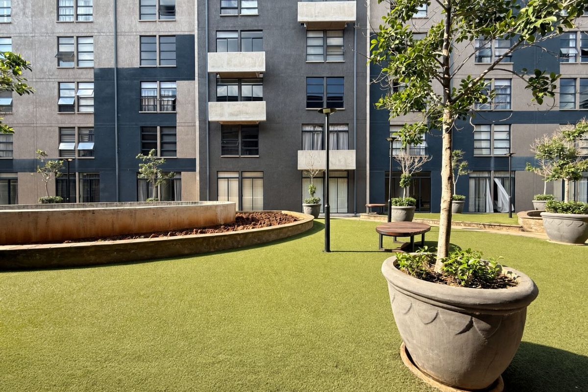 A well-maintained communal courtyard area featuring artificial turf, raised concrete planters, and exterior lighting, set against a modern multi-story apartment building facade.