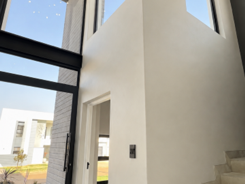 A bright, double-volume entrance hall featuring modern tiled stairs, large floor-to-ceiling windows, and a contemporary chandelier, creating an airy and sophisticated welcome.