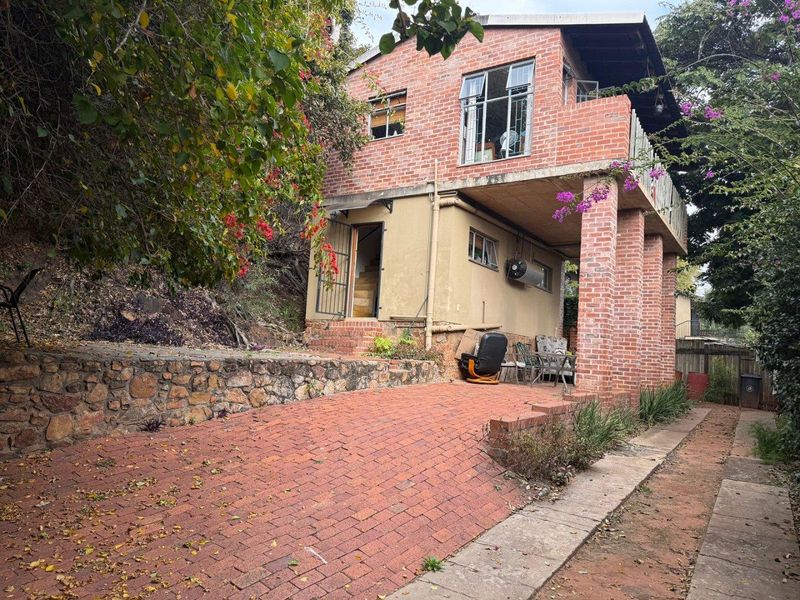 A two-story brick cottage featuring a paved driveway, stone retaining wall, and an elevated balcony area with exposed brick pillars.