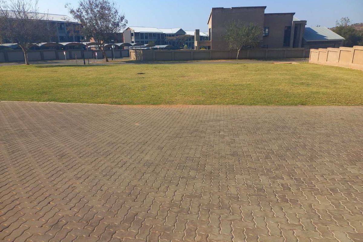 A view of a paved driveway leading to a spacious lawn area enclosed by a boundary wall, with neighboring buildings in the background.