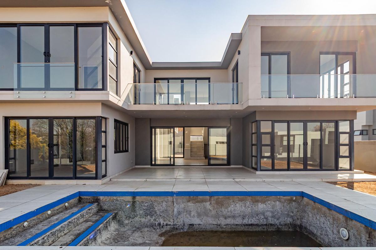 Rear view of a modern two-story house featuring extensive glass sliding doors, upper-level balconies with glass balustrades, and an unfinished swimming pool.