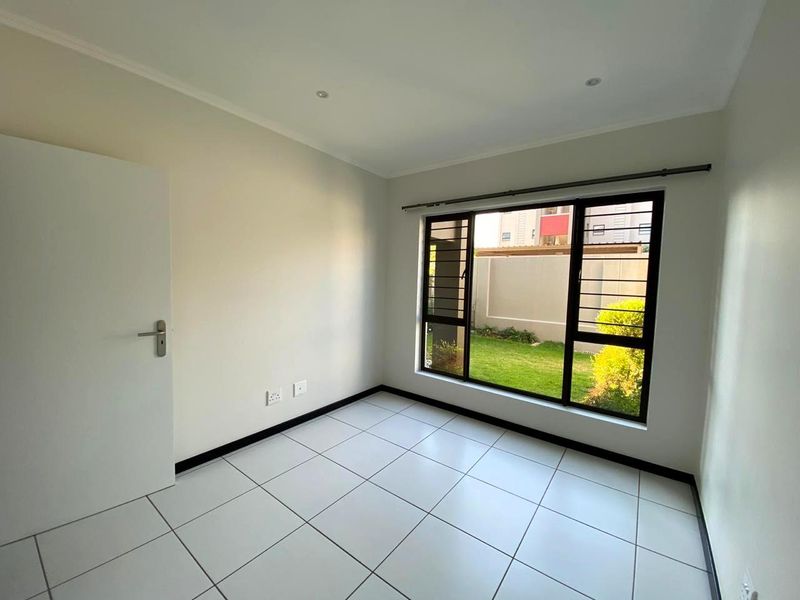 A bright, empty bedroom featuring neutral tiled flooring, recessed lighting, and a large window with security bars overlooking a private garden area.