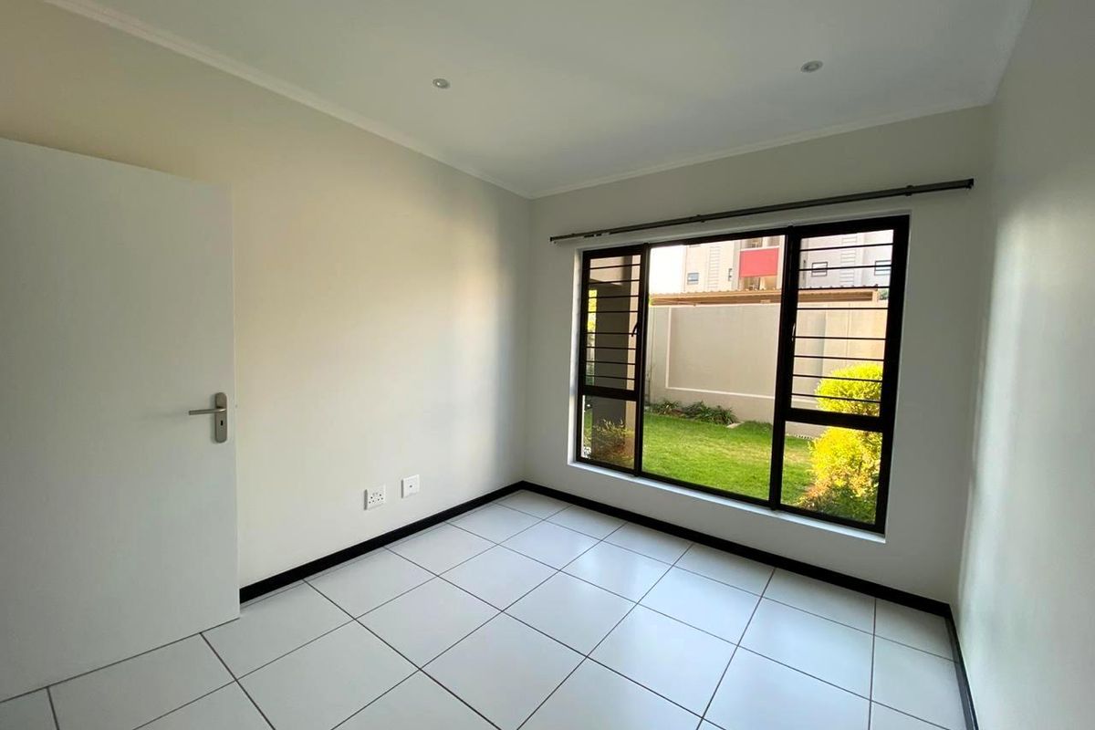 A bright, empty bedroom featuring neutral tiled flooring, recessed lighting, and a large window with security bars overlooking a private garden area.