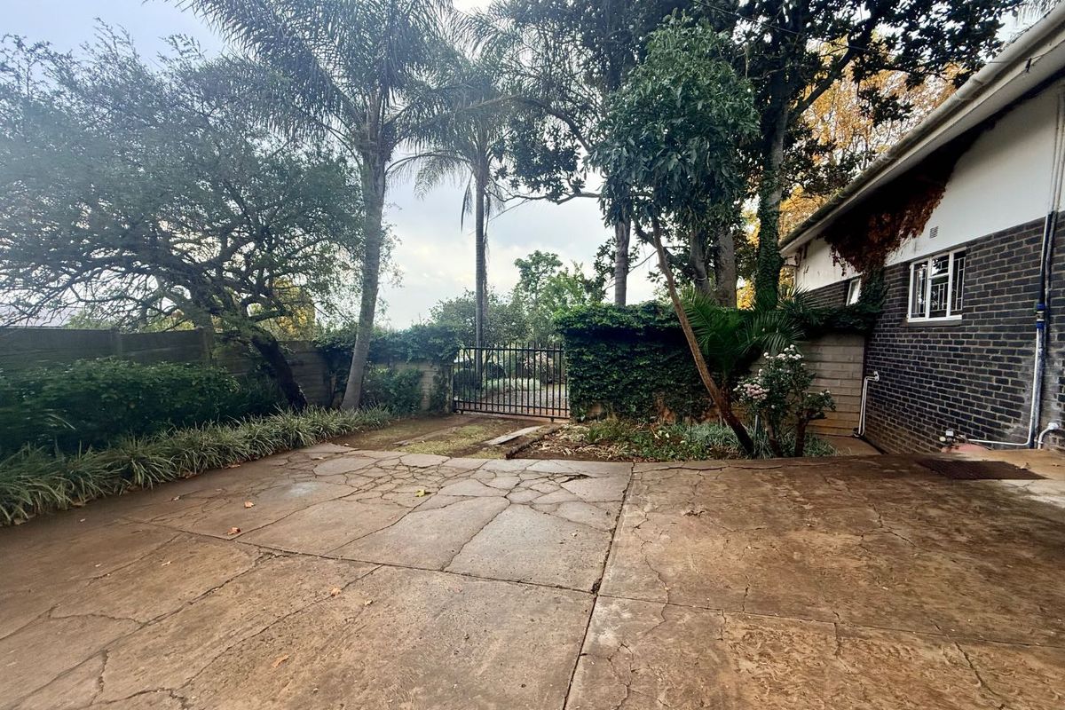 A rear exterior view of the property featuring a large, cracked concrete patio area, mature trees, and a metal security gate.