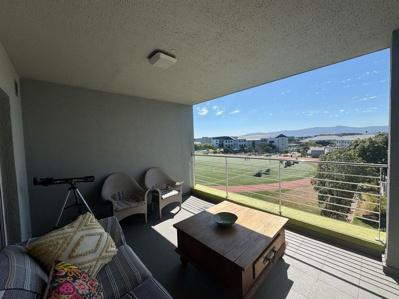 A spacious covered balcony featuring tiled flooring and a metal balustrade, offering an open view of the surrounding sports field and landscape.