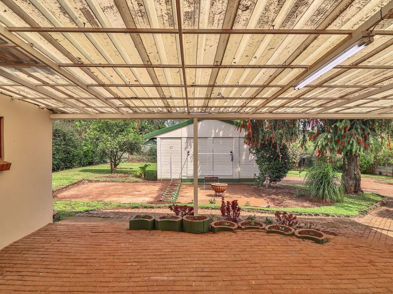 A covered outdoor patio area featuring brick paving and a corrugated roof, overlooking a spacious garden with a detached outbuilding.
