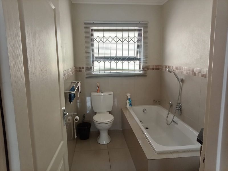 A functional bathroom featuring a built-in bathtub, toilet, and neutral wall tiling with a window fitted with security bars.