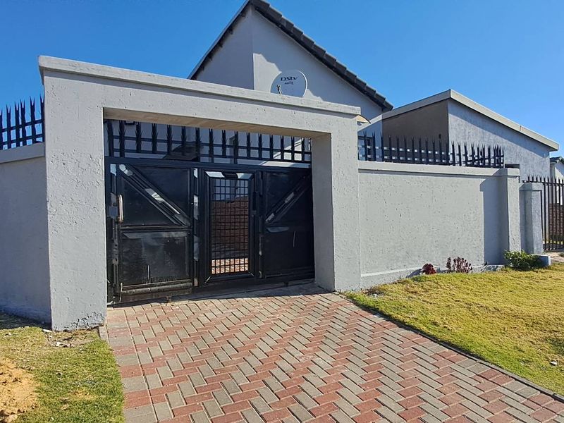 Front exterior view of the property featuring a secure perimeter wall with metal gates, paved driveway, and established lawn area.