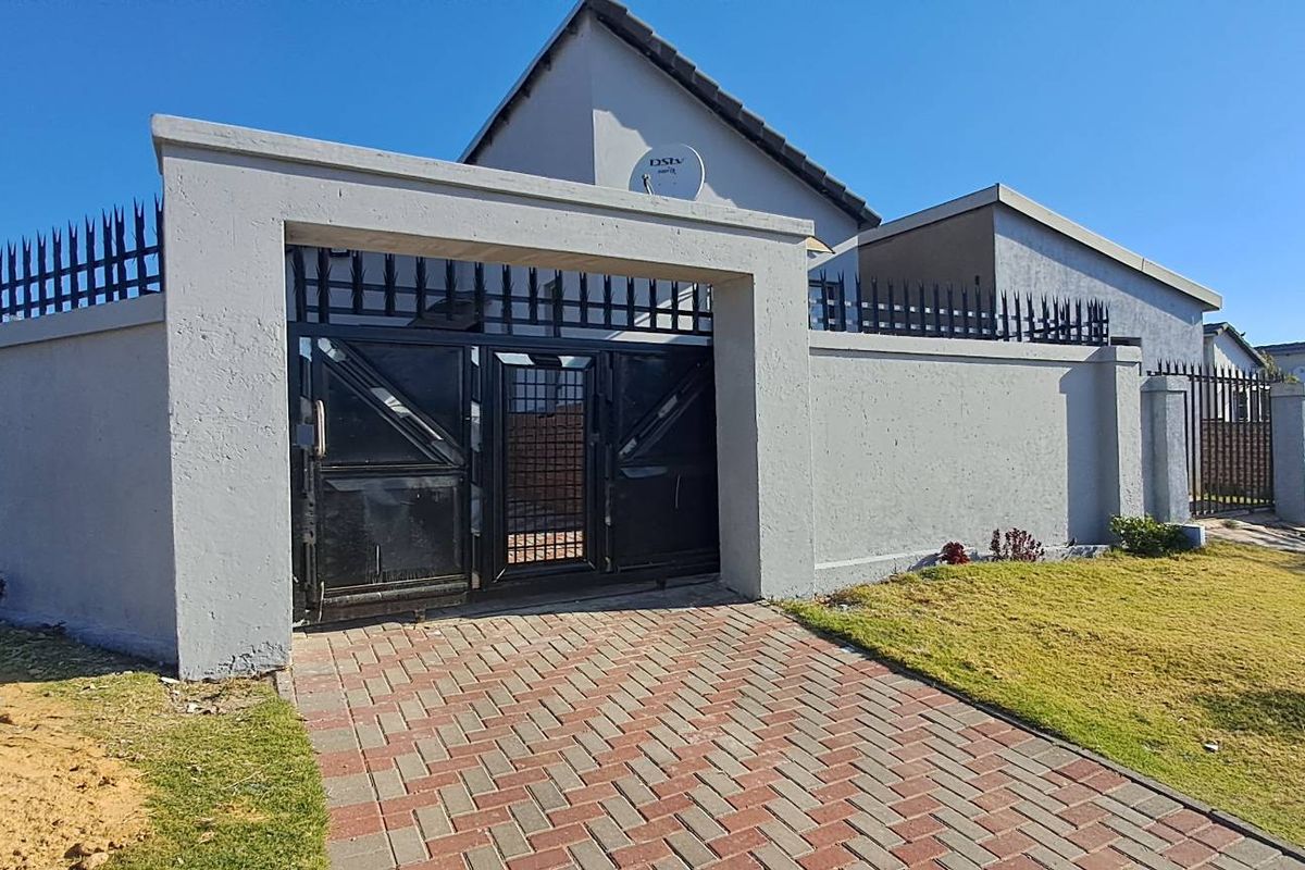 Front exterior view of the property featuring a secure perimeter wall with metal gates, paved driveway, and established lawn area.