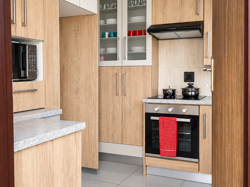 A modern, well-maintained kitchen featuring light wood-grain cabinetry, integrated appliances, sleek countertops, and polished tiled flooring, viewed through a doorway.