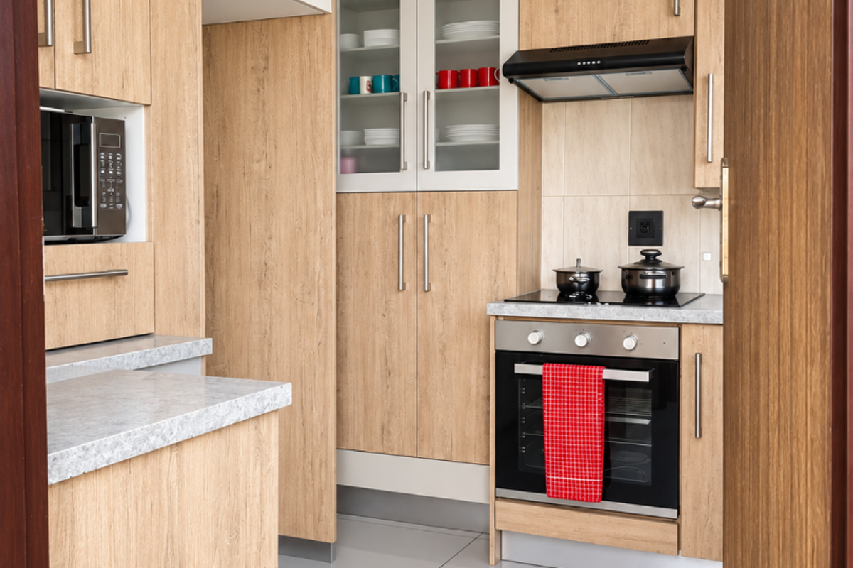 A modern, well-maintained kitchen featuring light wood-grain cabinetry, integrated appliances, sleek countertops, and polished tiled flooring, viewed through a doorway.