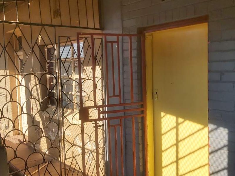 An exterior entrance area featuring a security gate, painted brick walls, and a concrete floor, showing signs of wear and requiring maintenance.