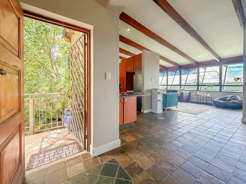 Open-plan living area featuring exposed timber beams, slate tile flooring, and expansive windows with natural light, leading to an outdoor balcony.