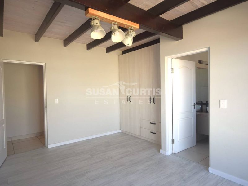 A bright, empty bedroom featuring exposed timber ceiling beams, built-in cupboards, and light-toned laminate flooring, with direct access to an en-suite bathroom.
