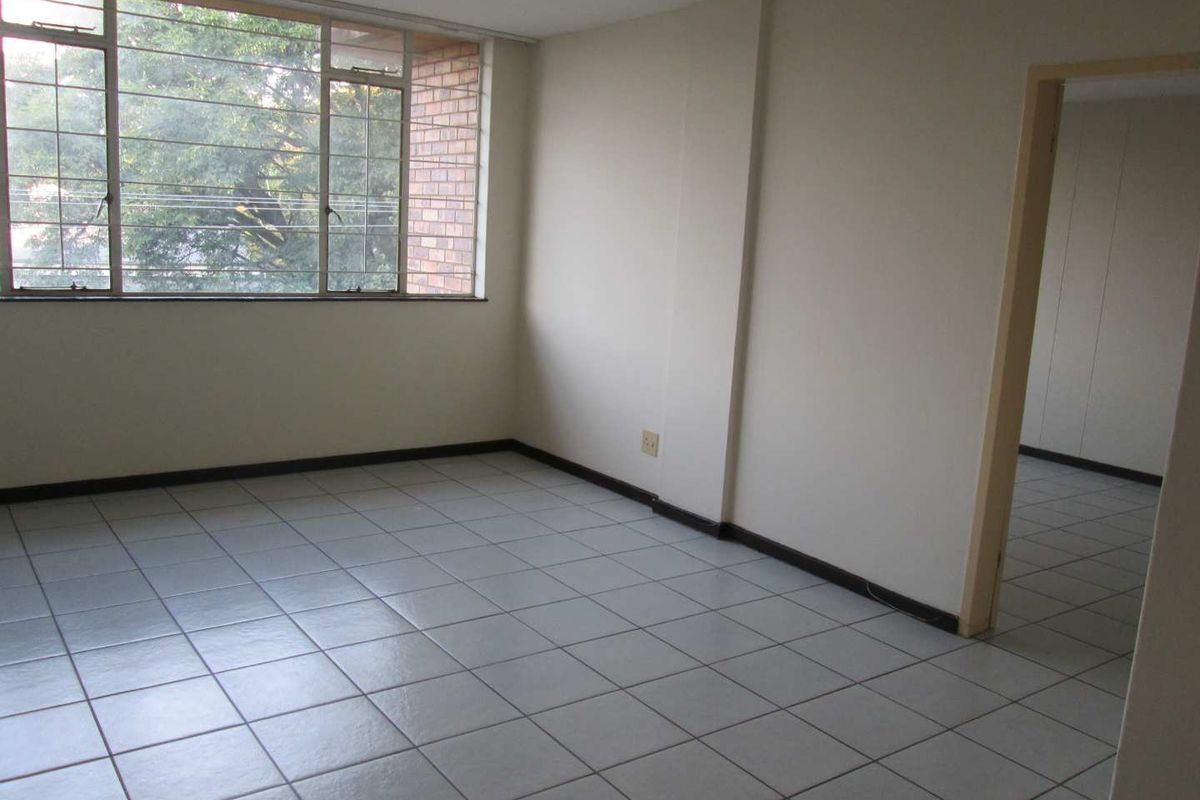 An empty, neutrally painted room featuring tiled flooring, large windows with metal frames, and a doorway leading to an adjacent space.
