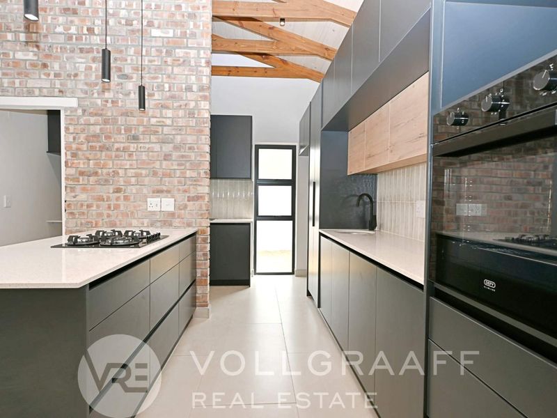 Modern kitchen featuring exposed brickwork, timber ceiling beams, sleek dark cabinetry, stone countertops, and integrated appliances in a contemporary open-plan layout.