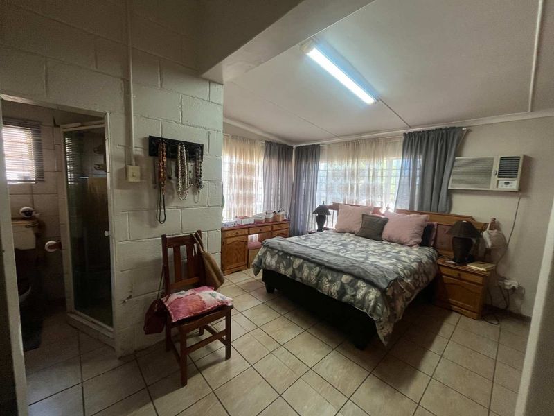 A bedroom featuring tiled flooring, an en-suite bathroom, and a window-mounted air conditioning unit, with exposed blockwork walls and dated finishes.