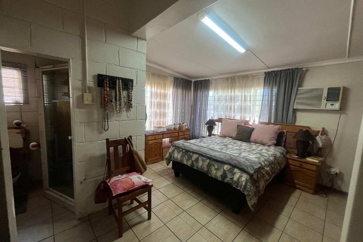 A bedroom featuring tiled flooring, an en-suite bathroom, and a window-mounted air conditioning unit, with exposed blockwork walls and dated finishes.