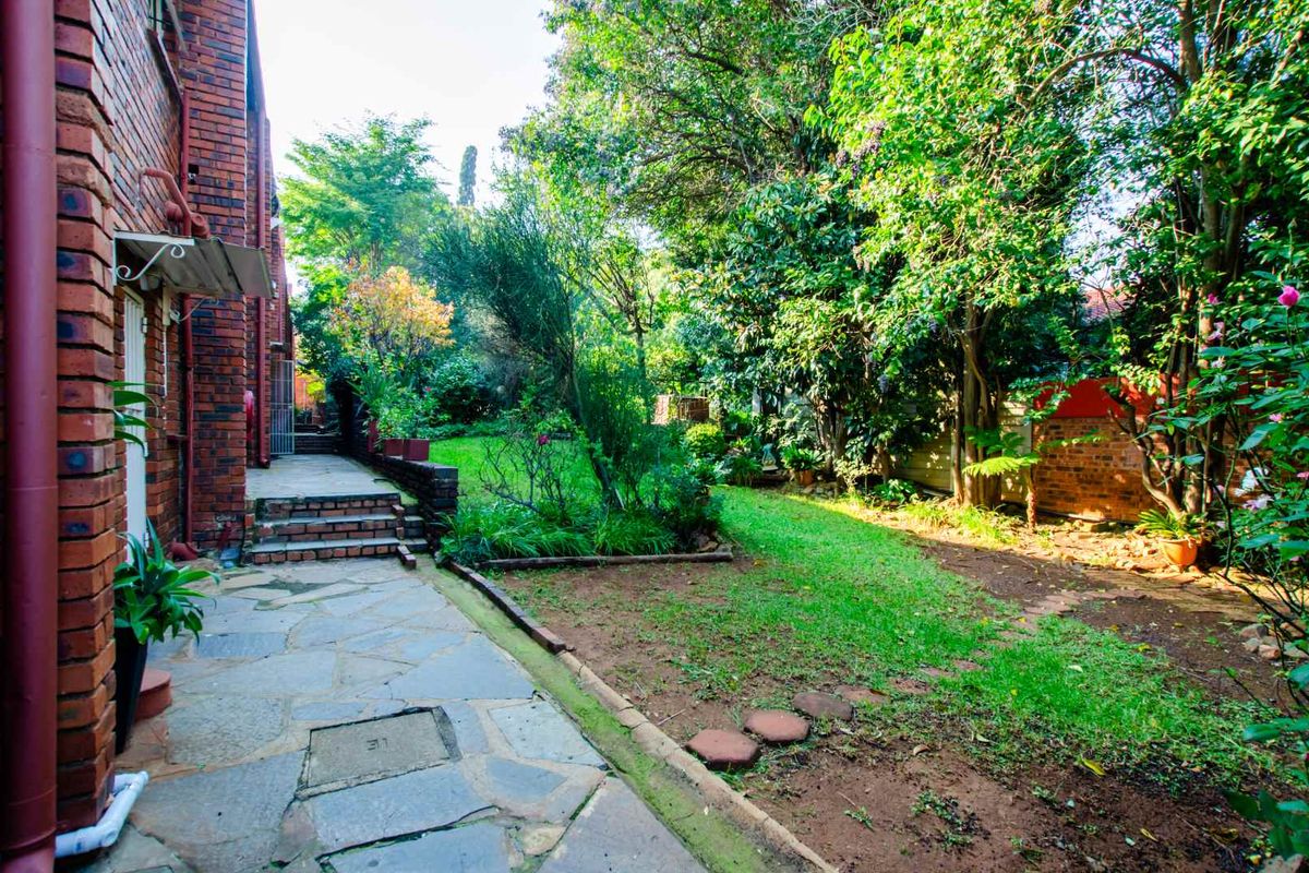 A private rear garden featuring a stone-paved walkway, established trees, and a lawn area adjacent to the brick building exterior.