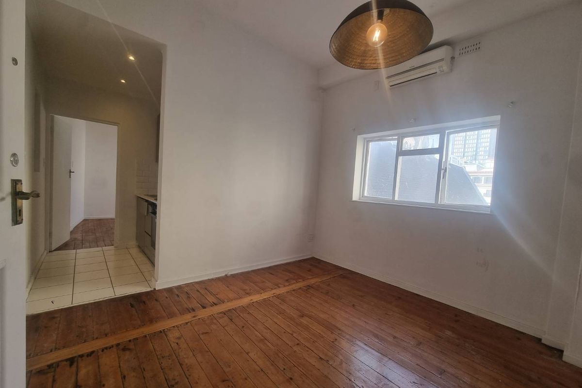 An empty living room featuring original hardwood flooring, a wall-mounted air conditioning unit, and an adjacent tiled hallway leading to the kitchen area.