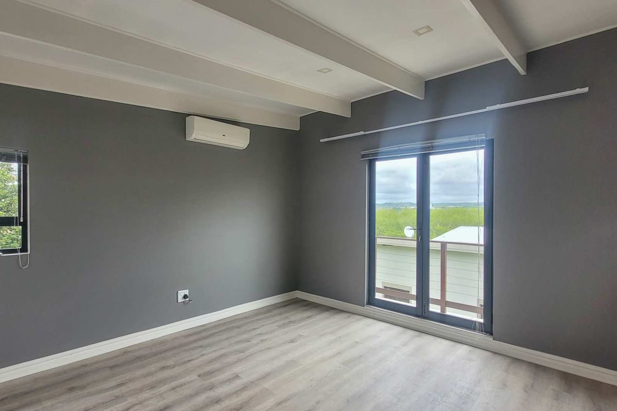 A bright, modern bedroom featuring grey walls, light wood-look flooring, exposed ceiling beams, a wall-mounted air conditioning unit, and glass doors leading outside.