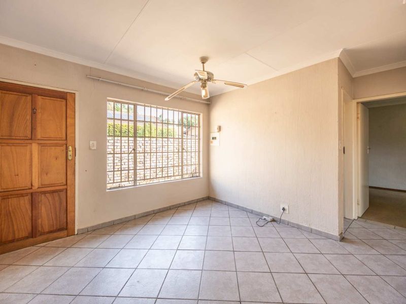 An empty living area featuring tiled flooring, a ceiling fan, and a wooden entrance door with security bars on the window.