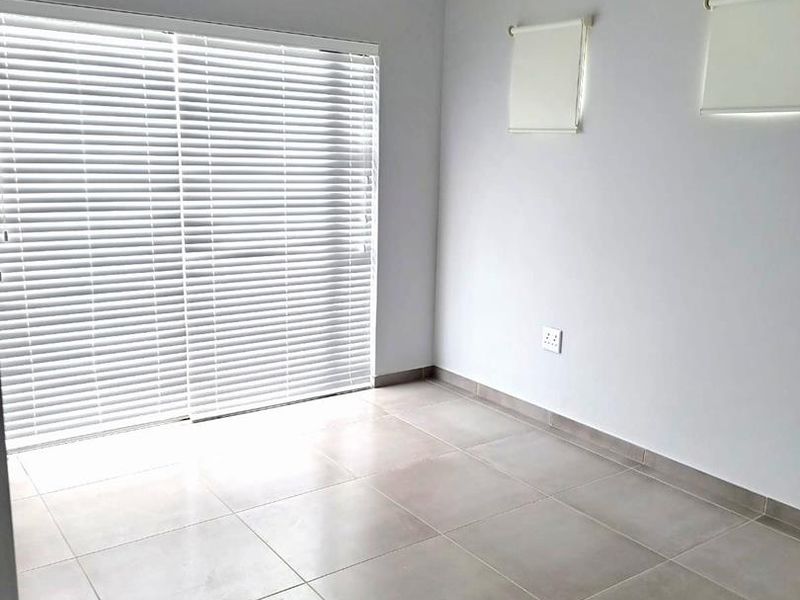 A bright, empty living area featuring neutral tiled flooring, recessed lighting, and a large sliding glass door with fitted venetian blinds.