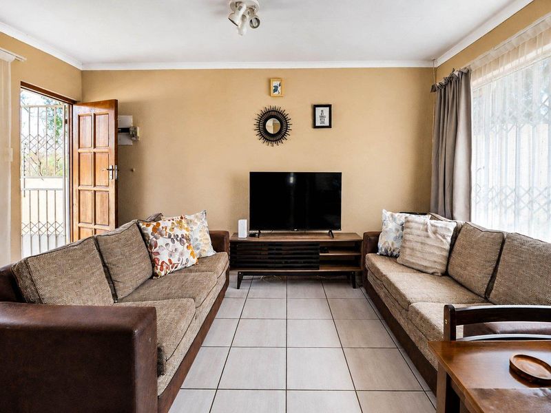 A comfortable living room featuring neutral tiled flooring, a wooden entrance door with security gate, and large windows allowing for natural light.