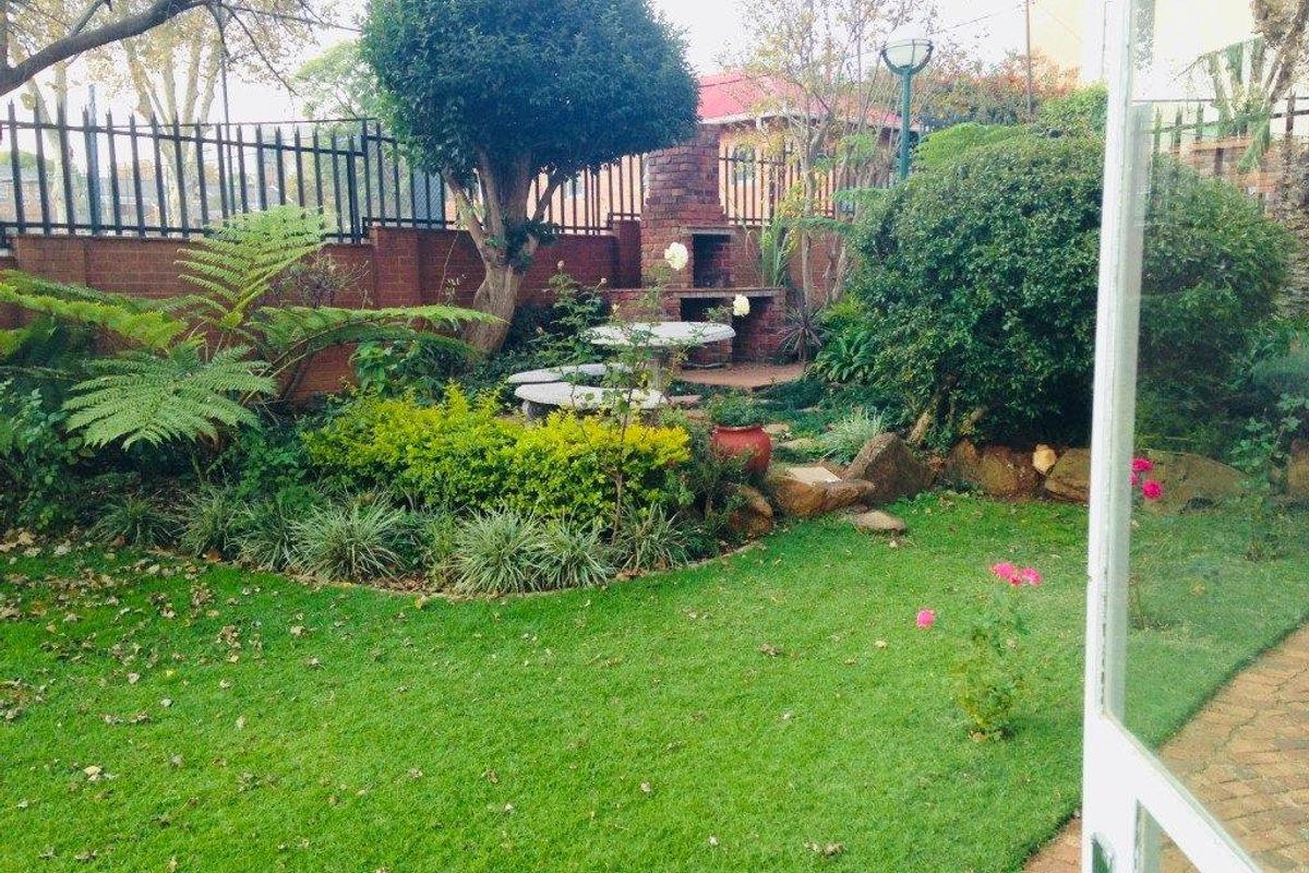 A private rear garden featuring a built-in brick braai area, established landscaping, and a lawn, viewed from the interior of the apartment.