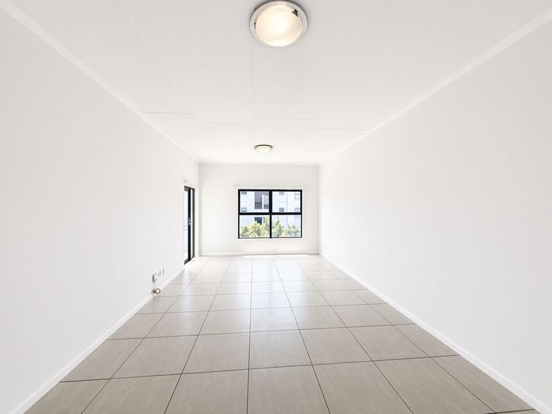 A bright, empty living area featuring neutral tiled flooring, clean white walls, and natural light from a large window and sliding glass door.