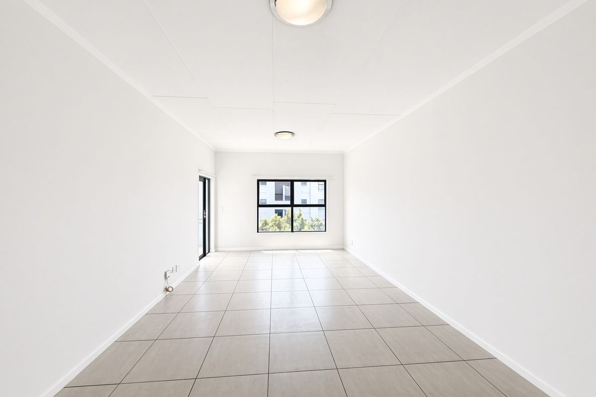 A bright, empty living area featuring neutral tiled flooring, clean white walls, and natural light from a large window and sliding glass door.
