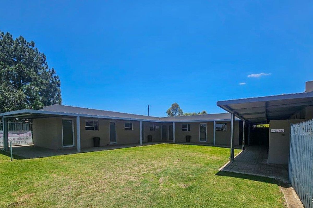 Rear view of the property featuring a covered patio area, paved walkways, and a spacious lawn, providing functional outdoor space for the building.