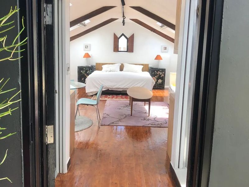 A spacious attic-style bedroom featuring exposed timber beams, polished hardwood flooring, and natural light from skylights, accessed through a distinctive arched doorway.