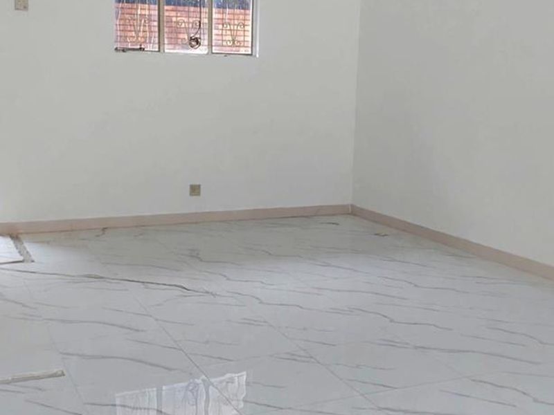 A bright, empty office space featuring modern marble-effect tiled flooring, a window with security bars, and a built-in air conditioning unit.