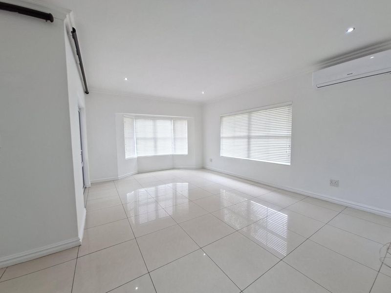 A bright, spacious, and empty living area featuring polished tiled floors, recessed lighting, a split-system air conditioner, and large windows with fitted venetian blinds.