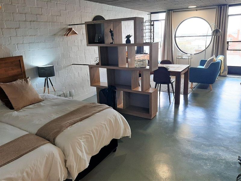 A modern open-plan studio apartment featuring a distinctive circular window, painted brick walls, and polished concrete flooring, creating a bright and industrial-style living space.