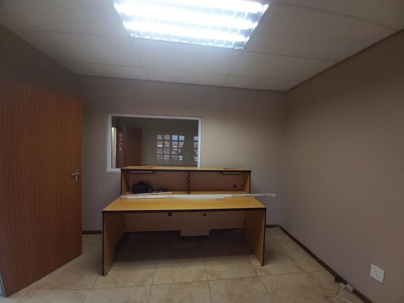 A functional office space featuring tiled flooring, a built-in reception desk, an internal window, and overhead fluorescent lighting.